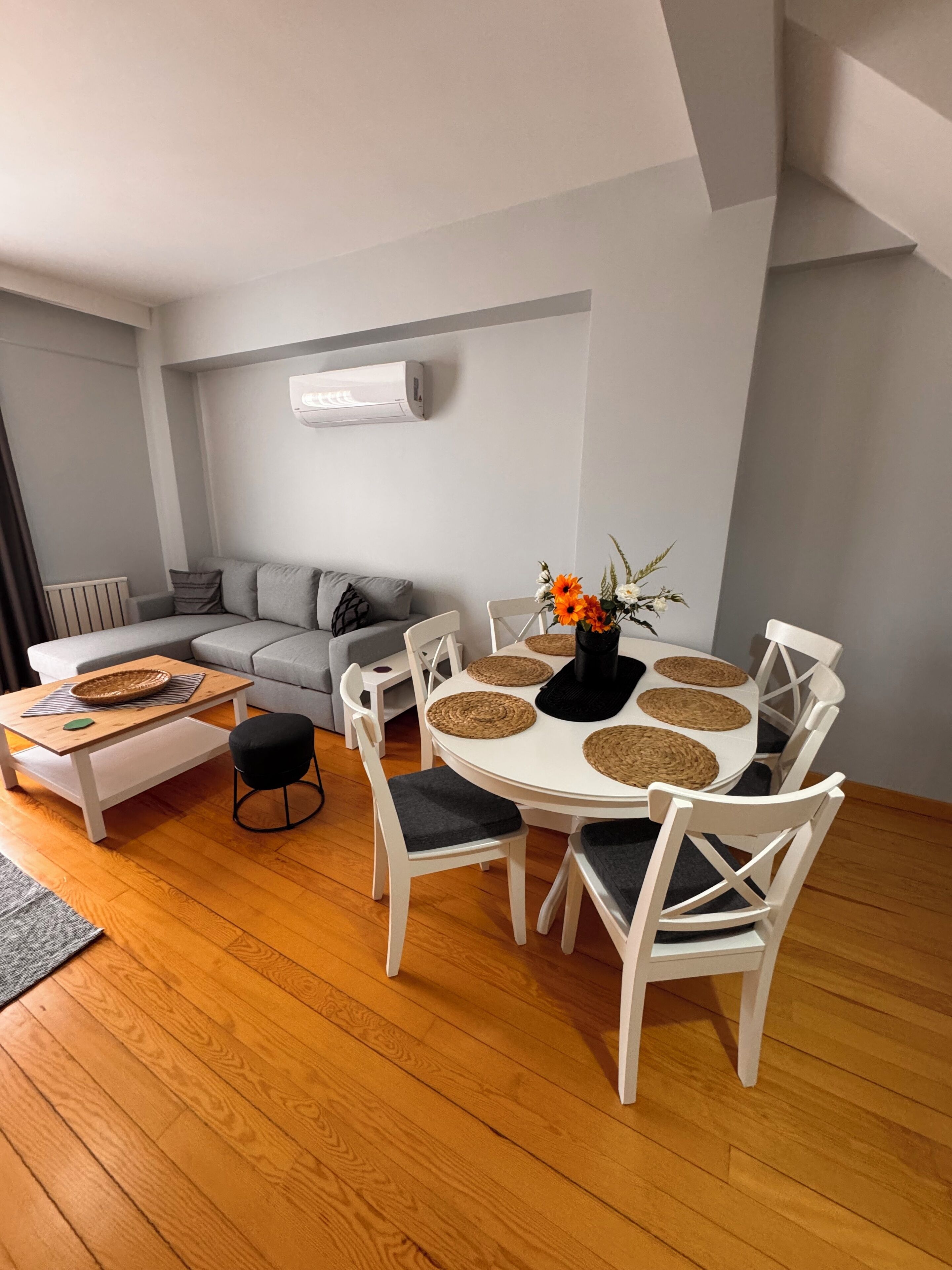 Un salon moderne avec un canapé gris, une table de salle à manger ronde entourée de chaises blanches et une table basse agrémentée d’un panier.