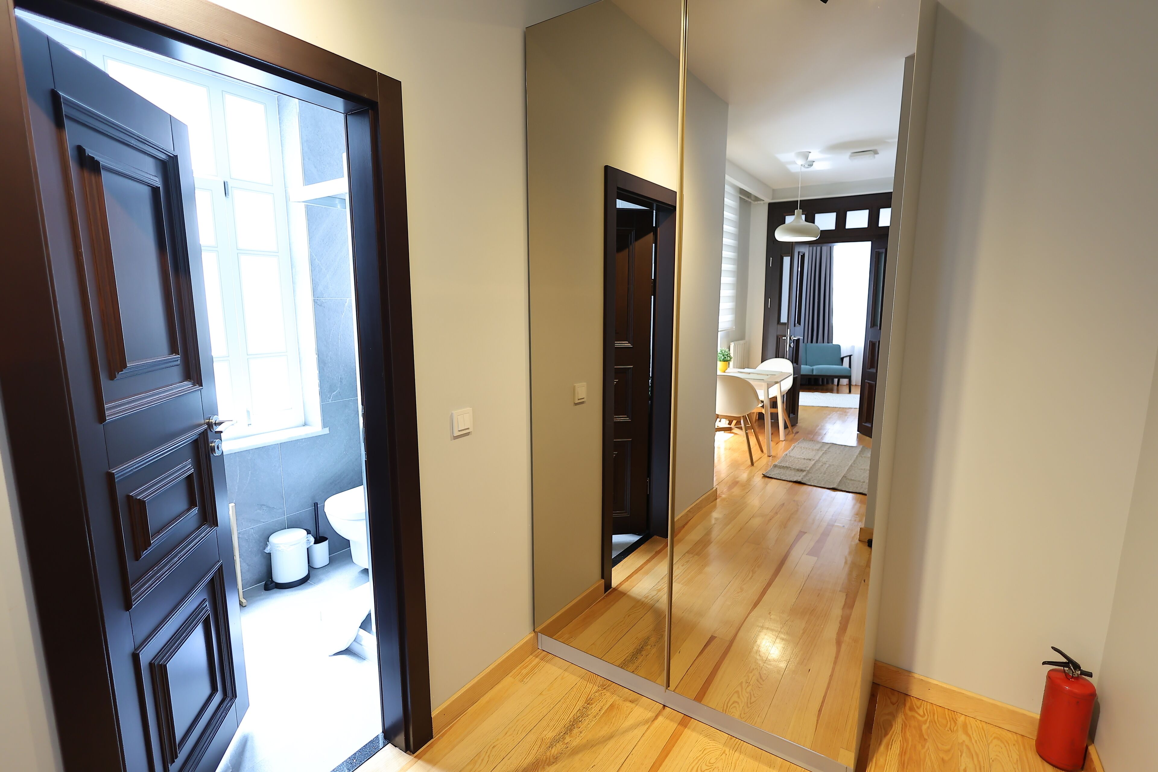 Un corridoio con pavimento in legno, una porta in legno scuro e un grande specchio che riflette una stanza con zona pranzo e una finestra.