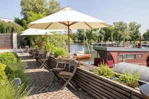 Un patio al aire libre con mobiliario de madera, una mesa con una cesta de pan y un barco al fondo.