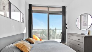 A modern bedroom with a large bed, a dresser, and a city view through a sliding door.