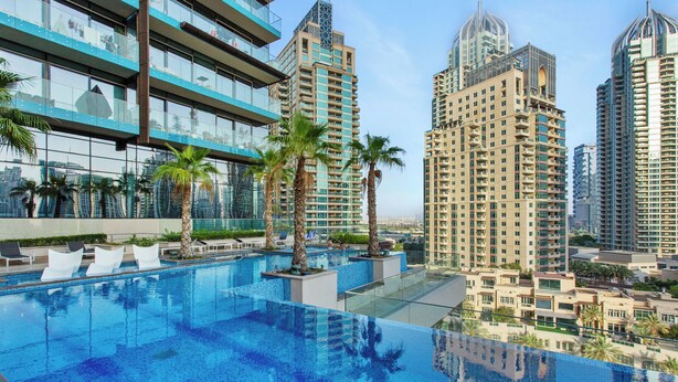 A rooftop infinity pool with clear blue water, surrounded by palm trees and modern buildings.