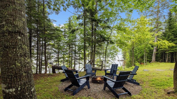 Une zone boisée avec un foyer et quatre chaises Adirondack disposées autour.