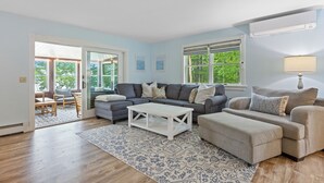 Un salon avec un canapé d’angle gris, une table basse blanche et un pouf beige. Un tapis à motifs recouvre le sol, et une fenêtre avec des stores se trouve sur le côté droit.