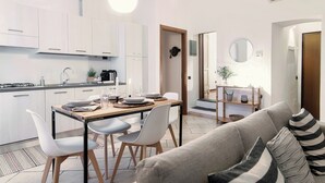 Et moderne køkken med hvide skabe, et spiseområde med et træbord og stole, samt en stue med sofa og spejl.
