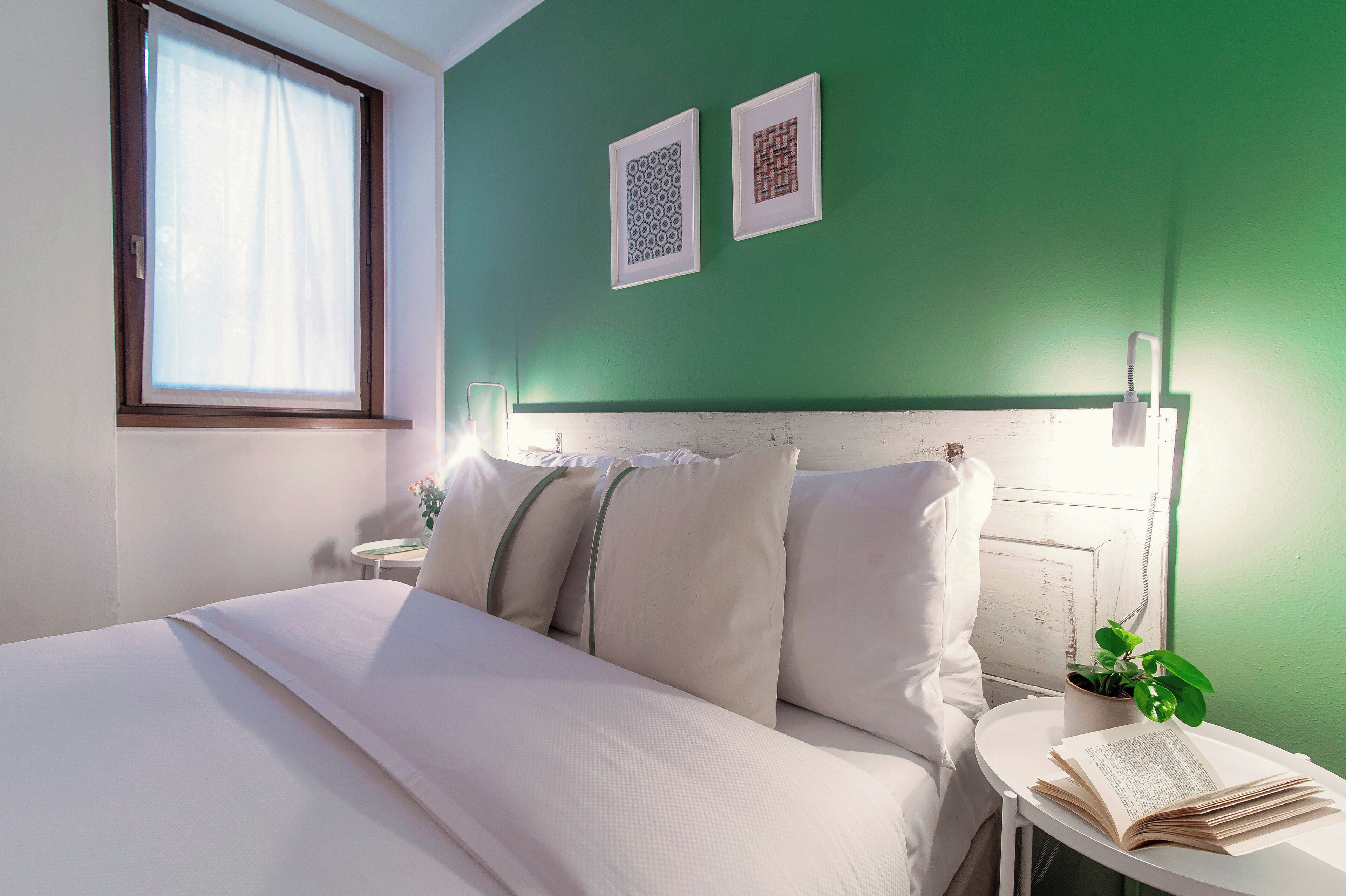 Una cama bien hecha con sábanas blancas, una mesita de noche con un libro y una planta en maceta, y dos cuadros en la pared.