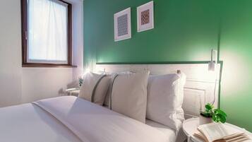 Una cama bien hecha con sábanas blancas, una mesita de noche con un libro y una planta en maceta, y dos cuadros en la pared.