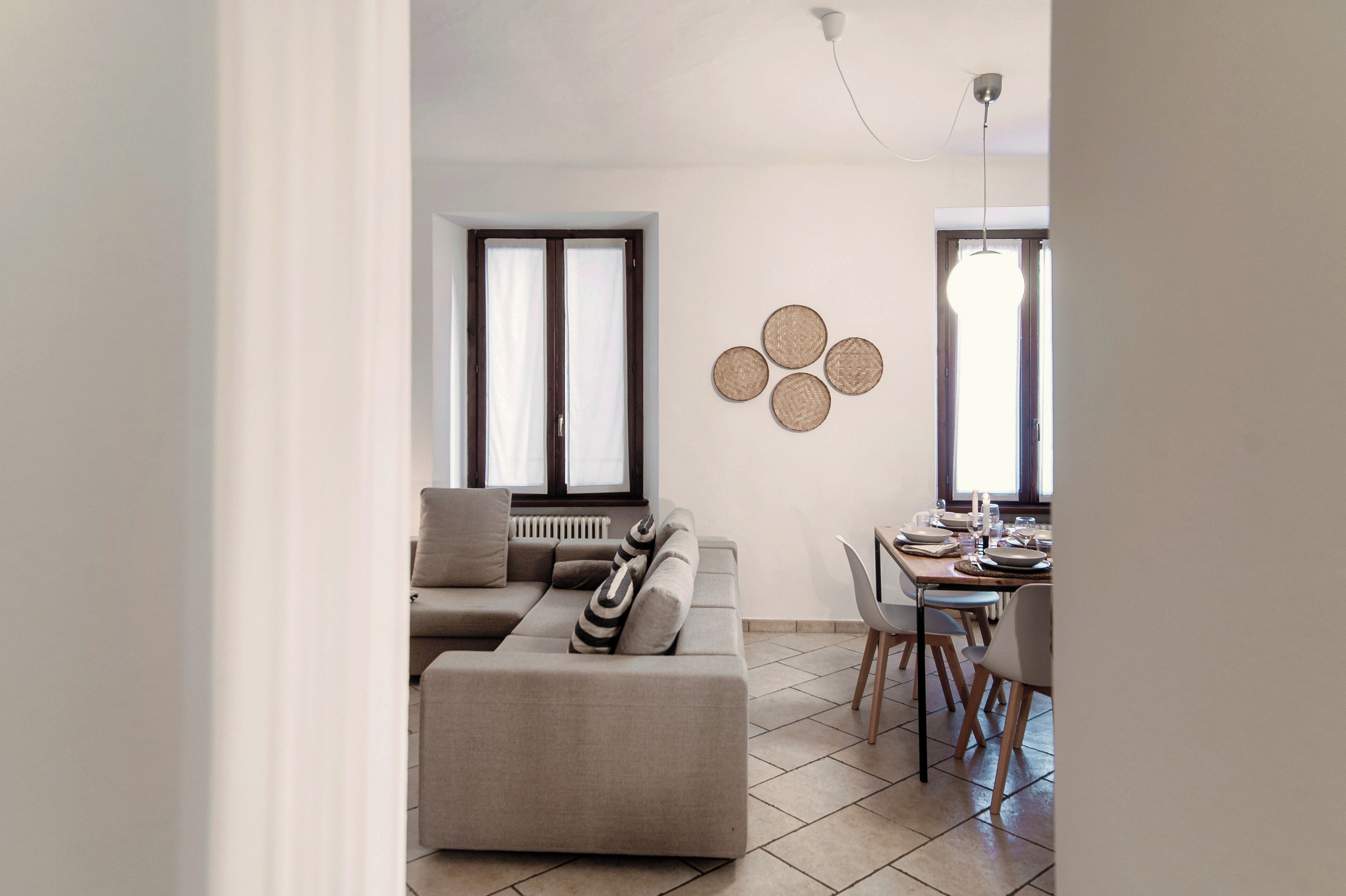 A modern living room with a sofa, dining table, and chairs. There are large windows with curtains, a radiator, and a wall-mounted light fixture.