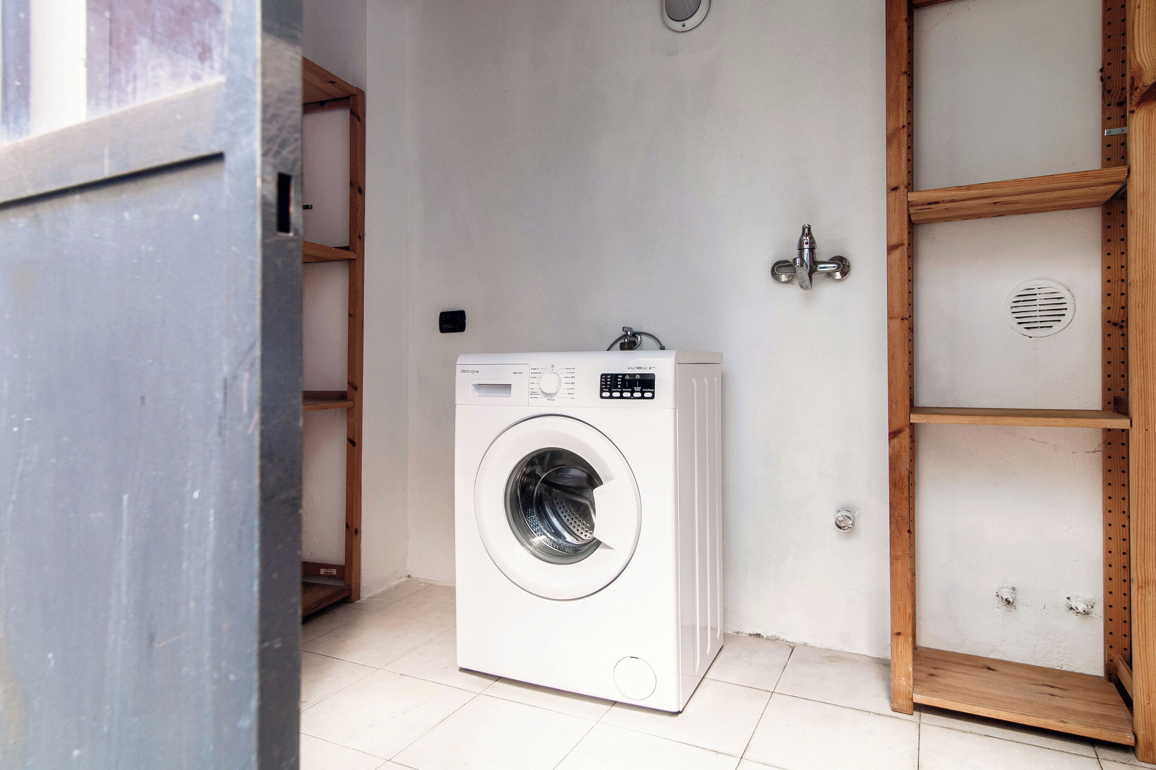 Eine kompakte, weiße Waschmaschine mit Frontlader in einem kleinen Waschraum mit einem Holzregal und einem Metalllüftungsgitter.