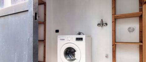A compact, white front-loading washing machine in a small laundry area with a wooden shelf and a metal vent.