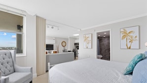Un dormitorio con una cama grande, un sillón gris, vistas a la ciudad y una cocina en el fondo.