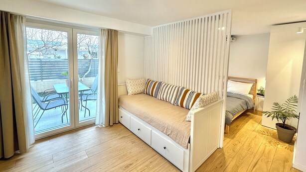 A compact room with a sofa bed, a small table, and a chair near a sliding glass door leading to an outdoor area.