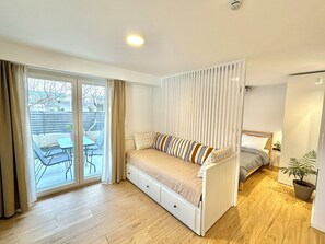 Een knus ingerichte kamer met een slaapbank, een klein tafeltje en een stoel naast een schuifdeur die uitkomt op een terras.