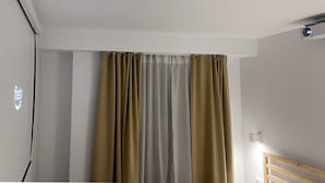 A room with a ceiling-mounted projector, a bed with a wooden headboard, and a window with beige curtains.