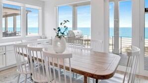 A dining area with a wooden table and white chairs, a white vase with blue flowers, and a view of the ocean through large windows. - Viviana #12 | Luxury Gulf Front Home, Private Pool + Incredible Views! (Pensacola)