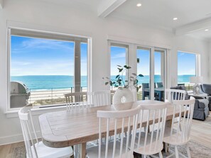 Comedor con mesa de madera y sillas blancas, ventanas amplias con vista al océano y un centro de mesa con plantas.