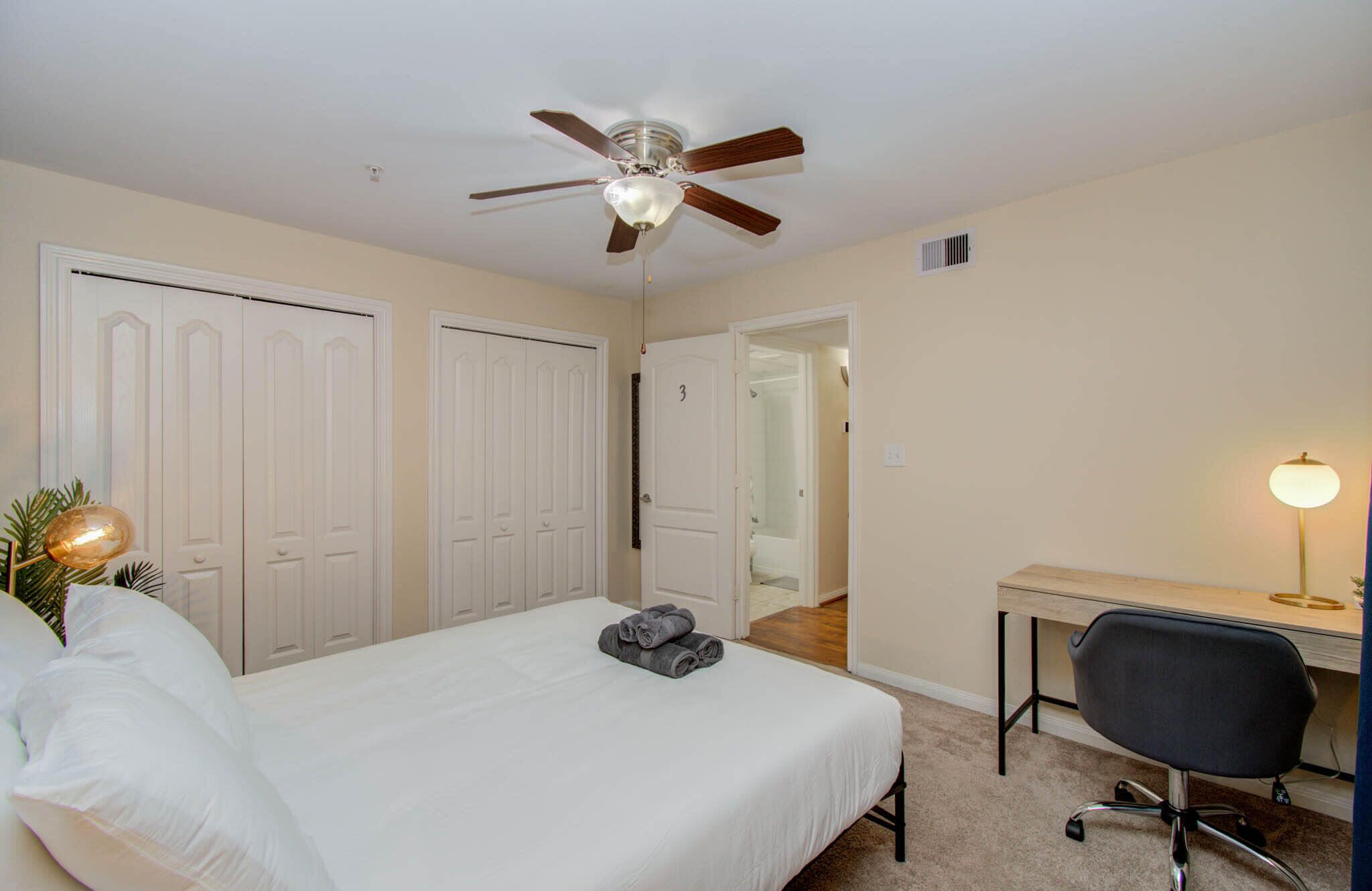 A hotel room with a bed, a desk, a chair, a ceiling fan, and a door leading to a bathroom.