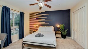 Un dormitorio con cama, ventilador de techo, mesita de noche con lámpara, planta en maceta y ventana con cortinas.