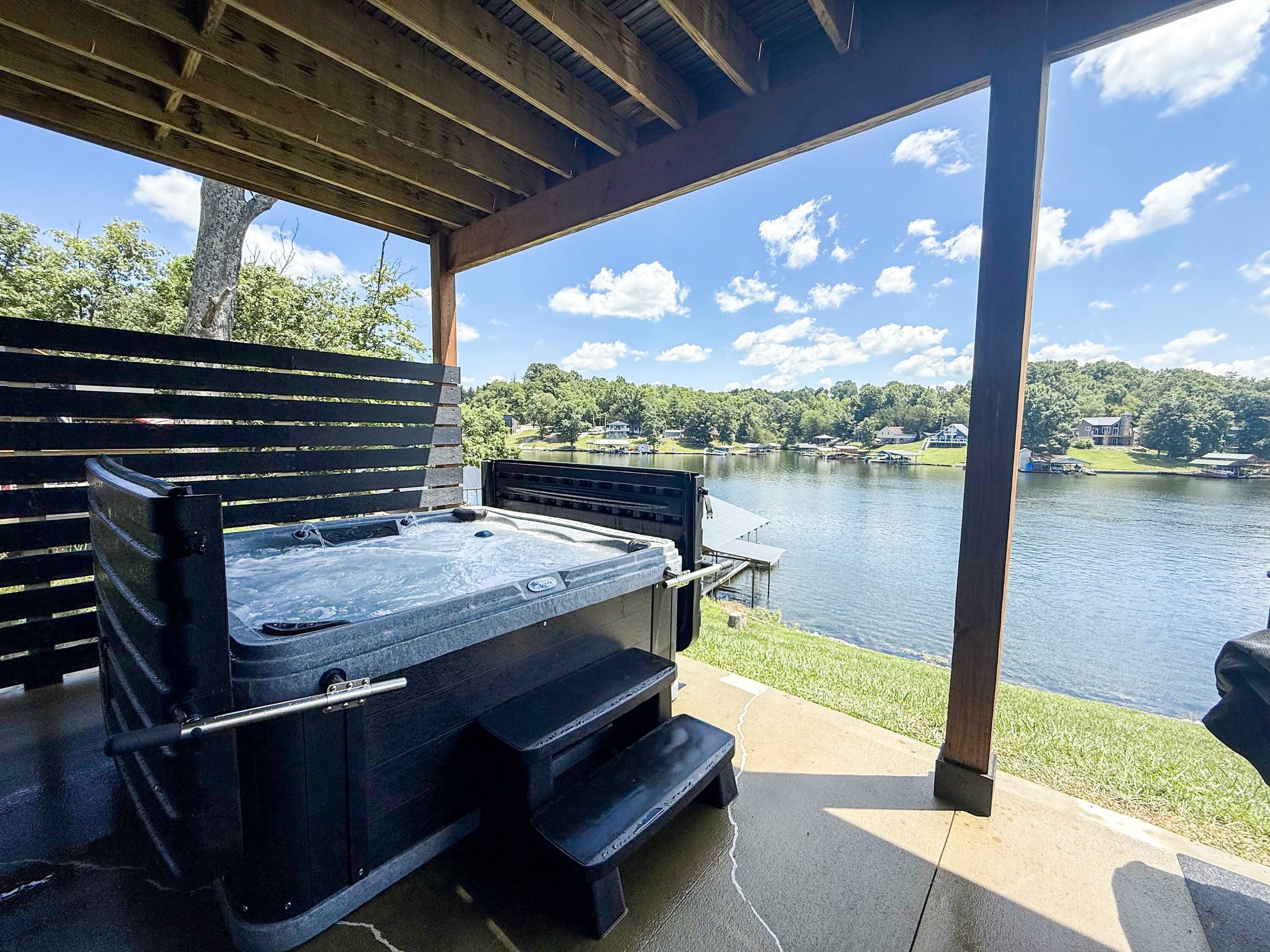 A hot tub under a wooden pergola with a view of a lake and houses in the background.