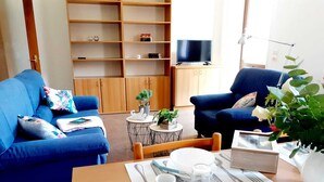 A living room with a blue sofa set, a wooden dining table with cutlery, a small round coffee table, and a TV on a cabinet. - Spazioso Appartamento Alle Porte Delle Dolomiti (Arten)