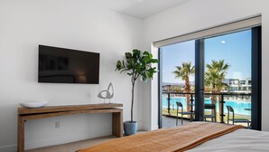 A modern hotel room with a large bed, a wooden desk, a television, a potted plant, and a balcony with a pool view. - Modern Lagoon: Desert Color Lagoon + Beaches (St. George)