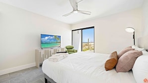 A modern bedroom with a bed, pillows, a TV, a chair, and a view of the beach.
