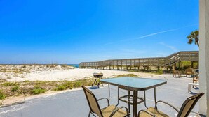A beachside patio with a table and chairs, a barbecue grill, and a wooden deck overlooking the beach.