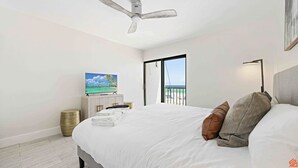 A hotel room with a bed, a ceiling fan, a TV, a bedside lamp, and a view of the beach through a sliding door.