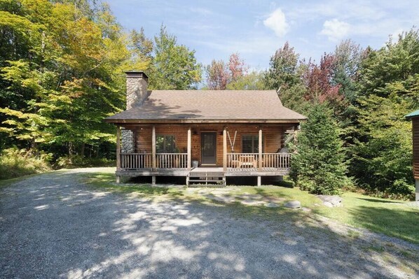 A log cabin with a stone chimney, a porch, and a gravel driveway surrounded by trees.