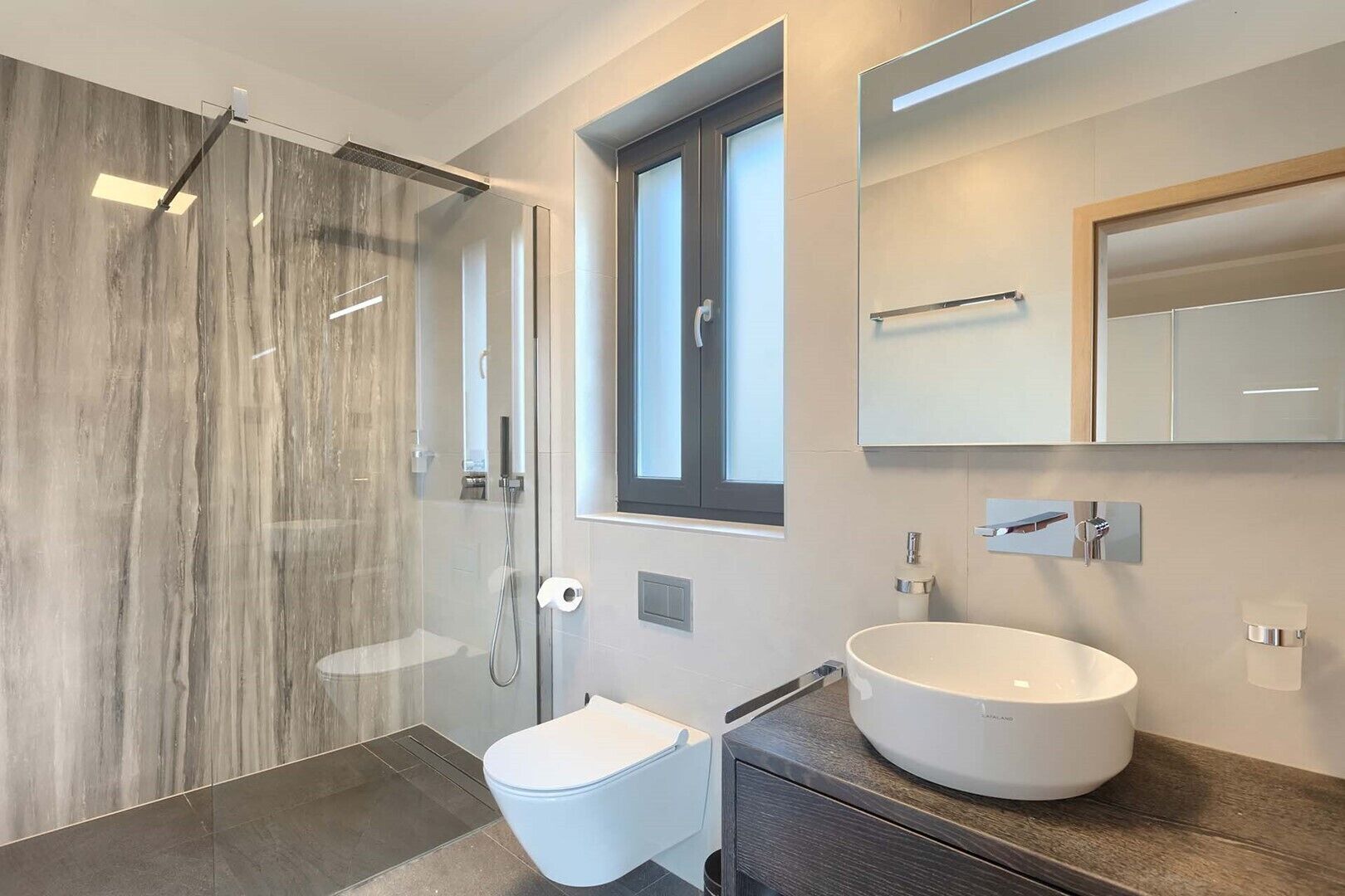 Une salle de bain moderne avec une douche à parois de verre, des toilettes blanches, un grand lavabo rond et un miroir au-dessus du lavabo.