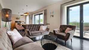 Sala de estar moderna com sofá, poltrona e mesa de centro. Há uma área de jantar com mesa e cadeiras, e uma cozinha ao fundo. O quarto tem janelas amplas e uma varanda com vista.