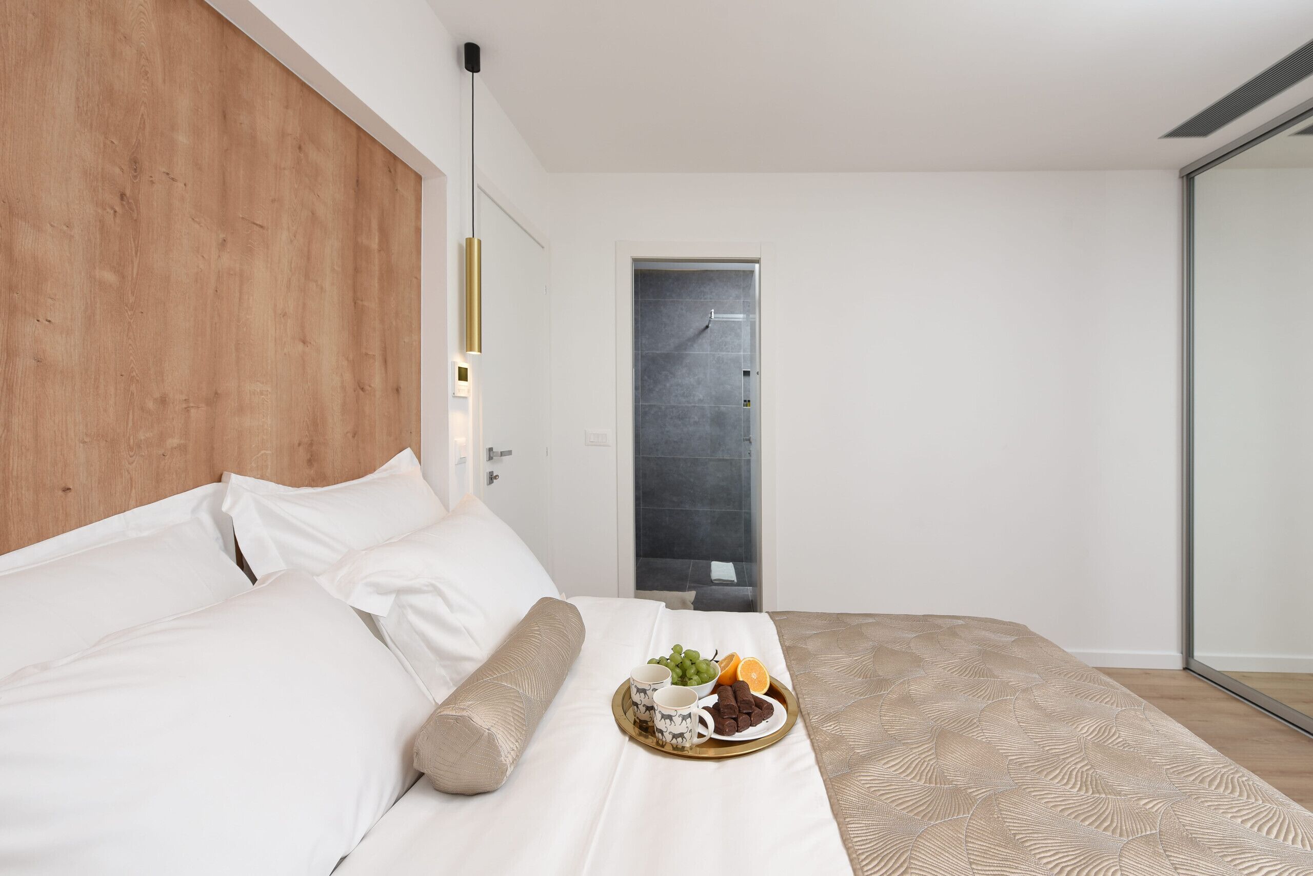 Un dormitorio moderno con cabecera de madera, cama con ropa de cama blanca, bandeja con frutas y bombones, y un espejo grande en la pared.
