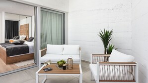 Ein modernes Hotelzimmer mit Bett, Sofa, kleinem Tisch mit Pflanze und Blick auf das danebenliegende Zimmer.
