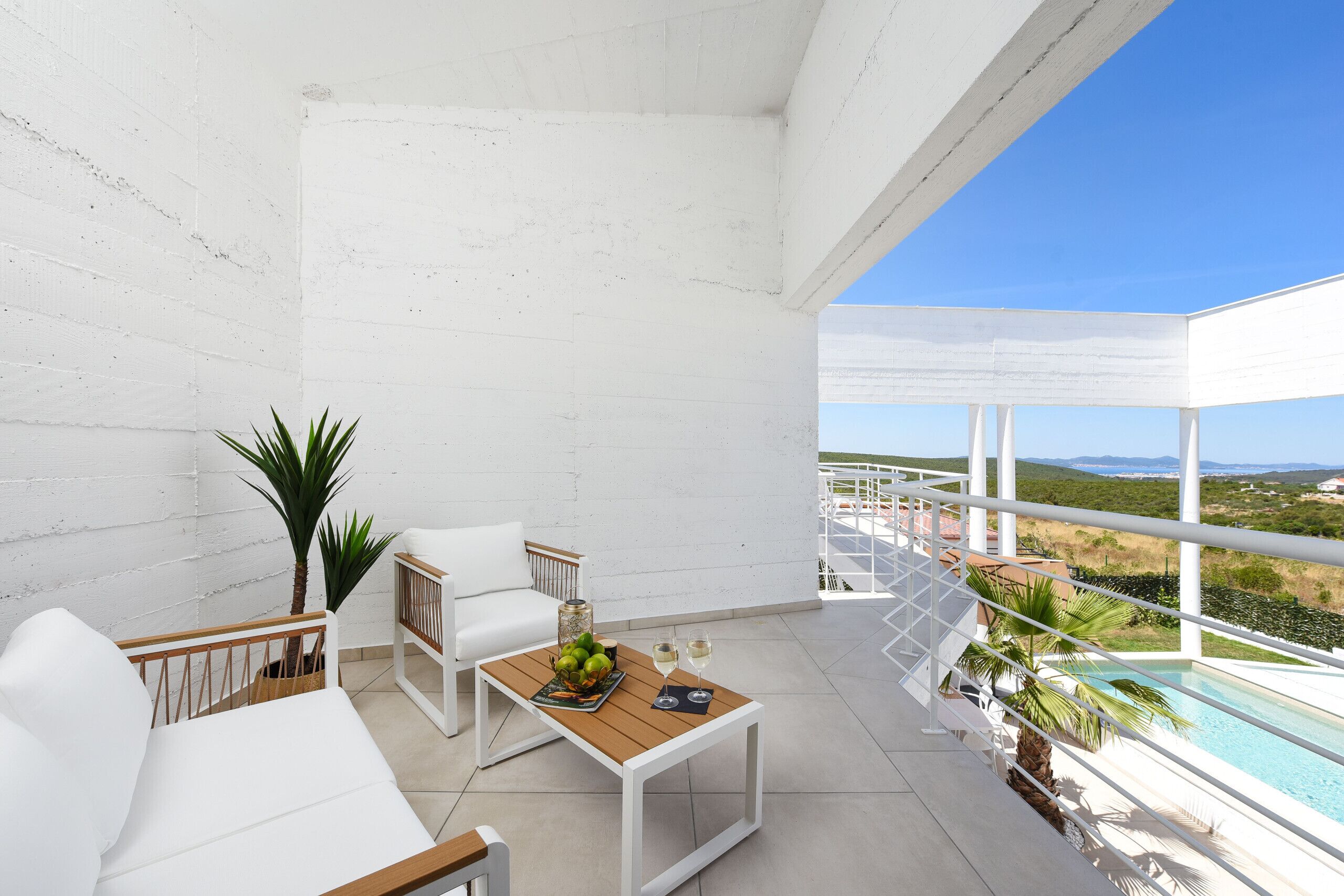 A balcony with white furniture, a wooden table with fruit and glasses, and a view of a pool and landscape.