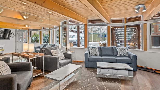 A modern living room with wooden beams, a grey sofa, a coffee table, and large windows.