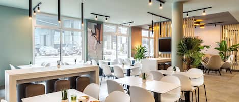 Un comedor moderno con mesas y sillas blancas, un ventanal con vistas a la calle y una zona de estar informal con plantas y un televisor.