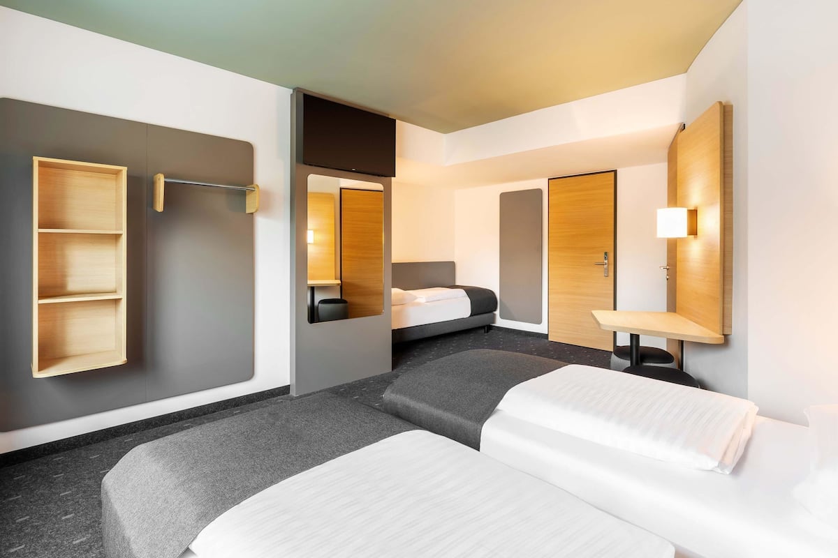 Una habitación de hotel moderna con alfombra gris, dos camas, un armario de madera, un espejo y una mesita de noche con lámpara.