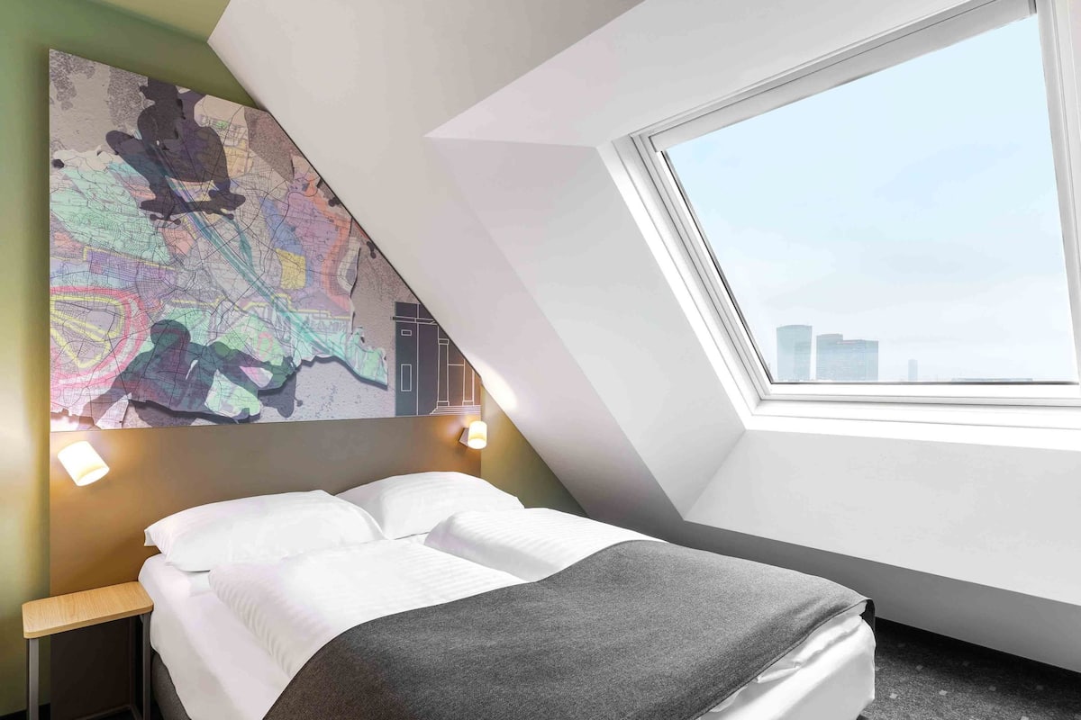 Una habitación de hotel moderna con una cama, una obra de arte con un mapa en la pared, un tragaluz y vistas a la ciudad.