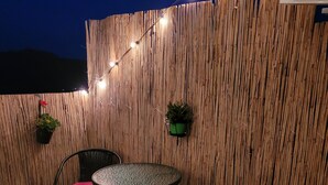 Un patio con pared de bambú, dos sillas con cojines rojos, una mesita redonda y luces colgantes.