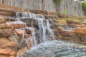 A waterfall cascading over rocks into a pool, surrounded by a wooden fence. - West End Lodge Resort Style Pool (Fredericksburg)