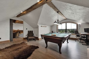 A spacious loft with a pool table, a large window offering a view, and a rustic wooden ceiling. - West End Lodge Resort Style Pool (Fredericksburg)