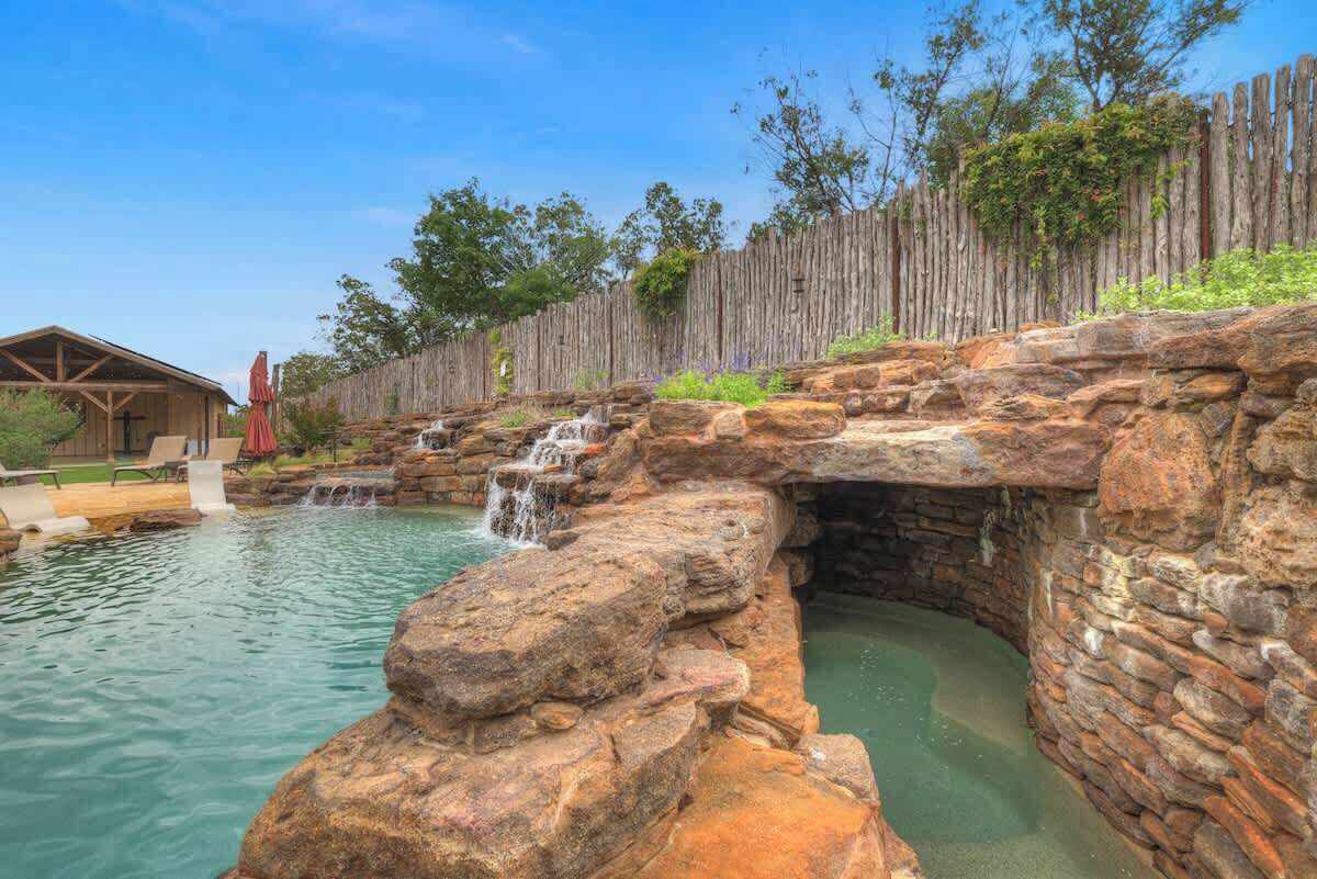 Une piscine d’aspect naturel, agrémentée d’une cascade, entourée d’une clôture en bois et bordée d’un bâtiment au style rustique.
