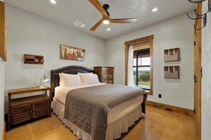 Une chambre à coucher avec un lit, une table de chevet, un ventilateur de plafond, une fenêtre et un tableau accroché au mur.