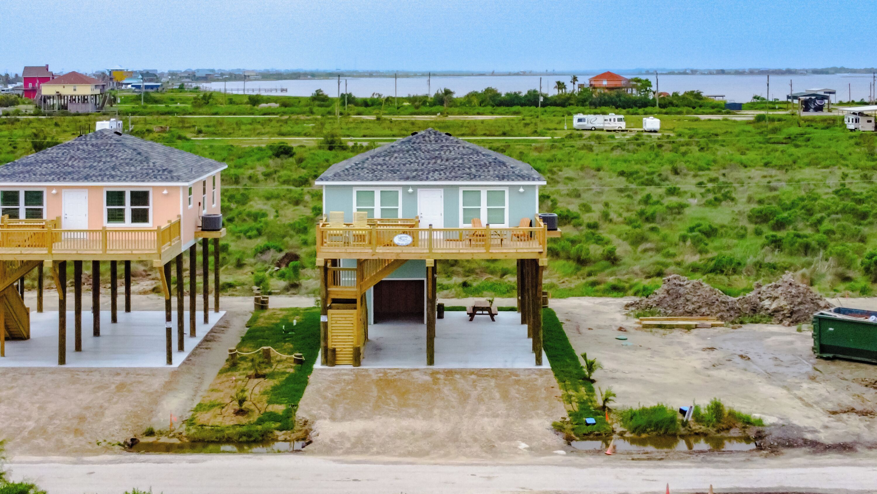 Deux maisons situées en bord de mer, dotées de terrasses surélevées et d’une zone sablonneuse devant.