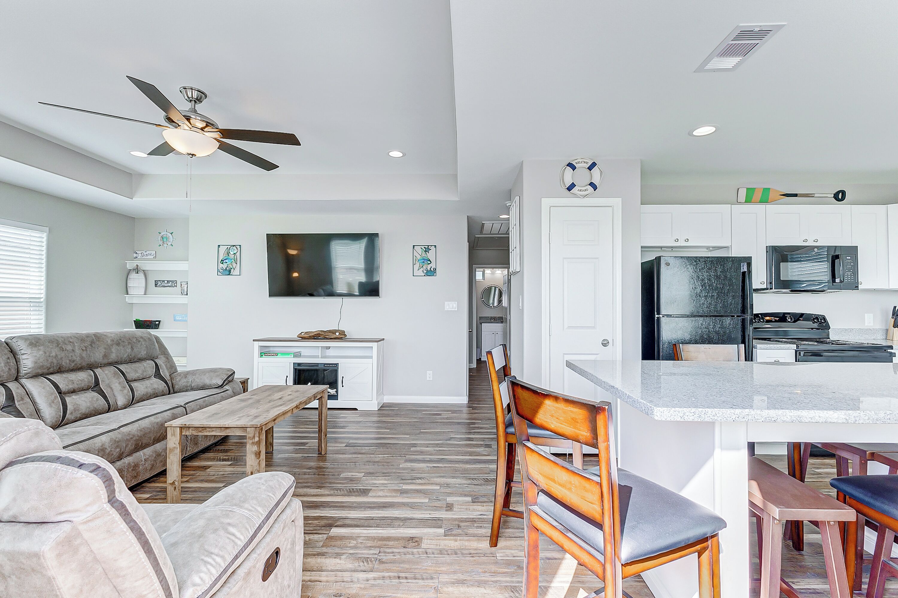 A modern living room with a sofa, coffee table, and chairs, a kitchen with white cabinets and appliances, and a ceiling fan.