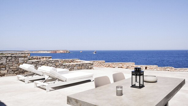 A terrace with white lounge chairs and a table, overlooking the sea with a stone wall and a clear sky. - Cave Suite Paros, 65sq.m | Private pool & Panoramic sea view | "endless blue"
 (Paros Cyclades Greece)