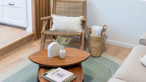 Ein Wohnzimmer mit rundem Holztisch, weißer Vase mit Blumen, Korbstuhl mit weißem Kissen, gewebtem Korb und einer Couch.