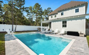 A residential swimming pool with a covered patio area, adjacent to a white house with a brown roof.