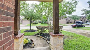 A covered porch with a brick wall, a stone pillar, and a view of a residential street with houses and cars.