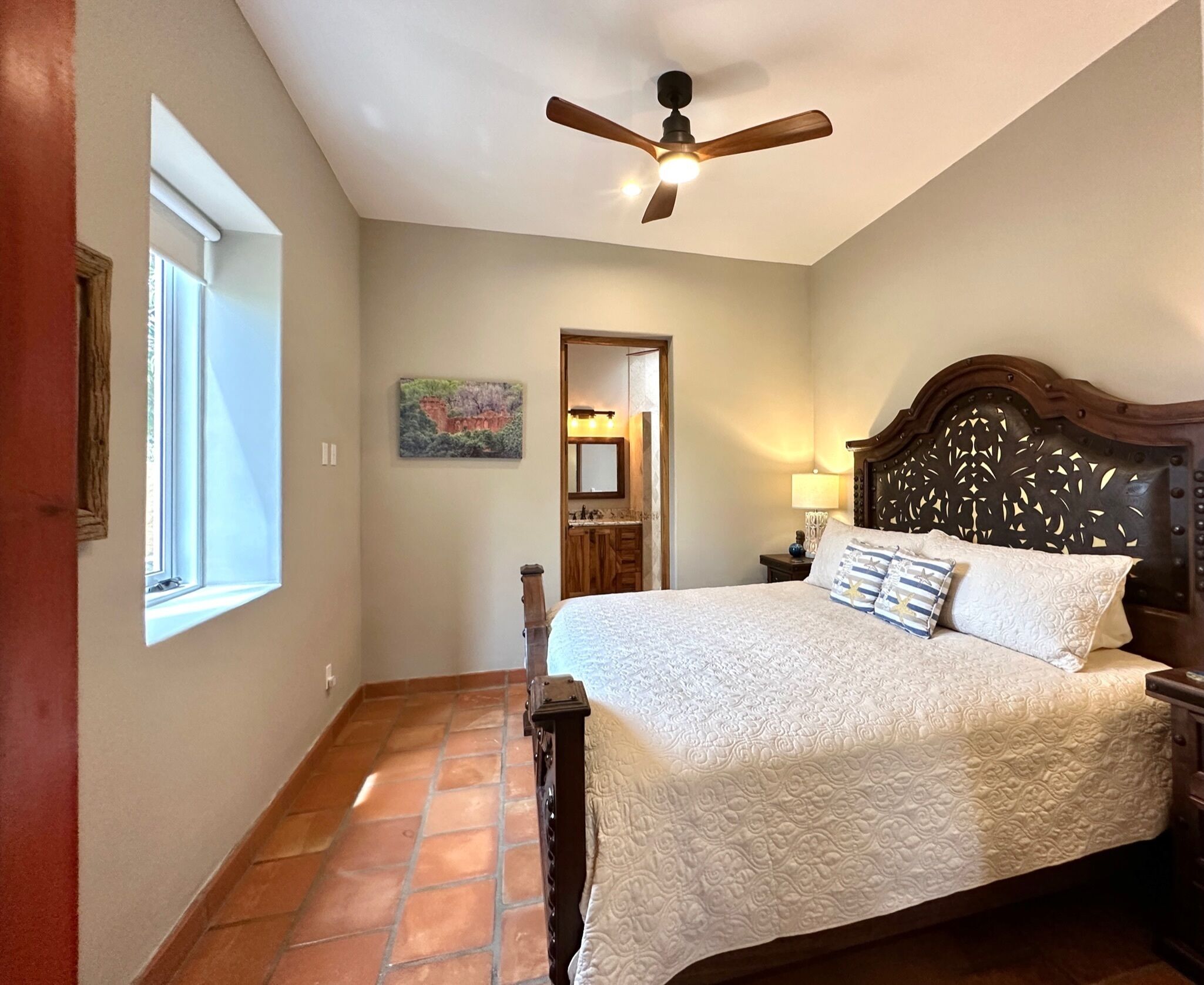 Une chambre à coucher avec un grand lit, un ventilateur de plafond, une fenêtre et une salle de bain visible à travers une porte ouverte.