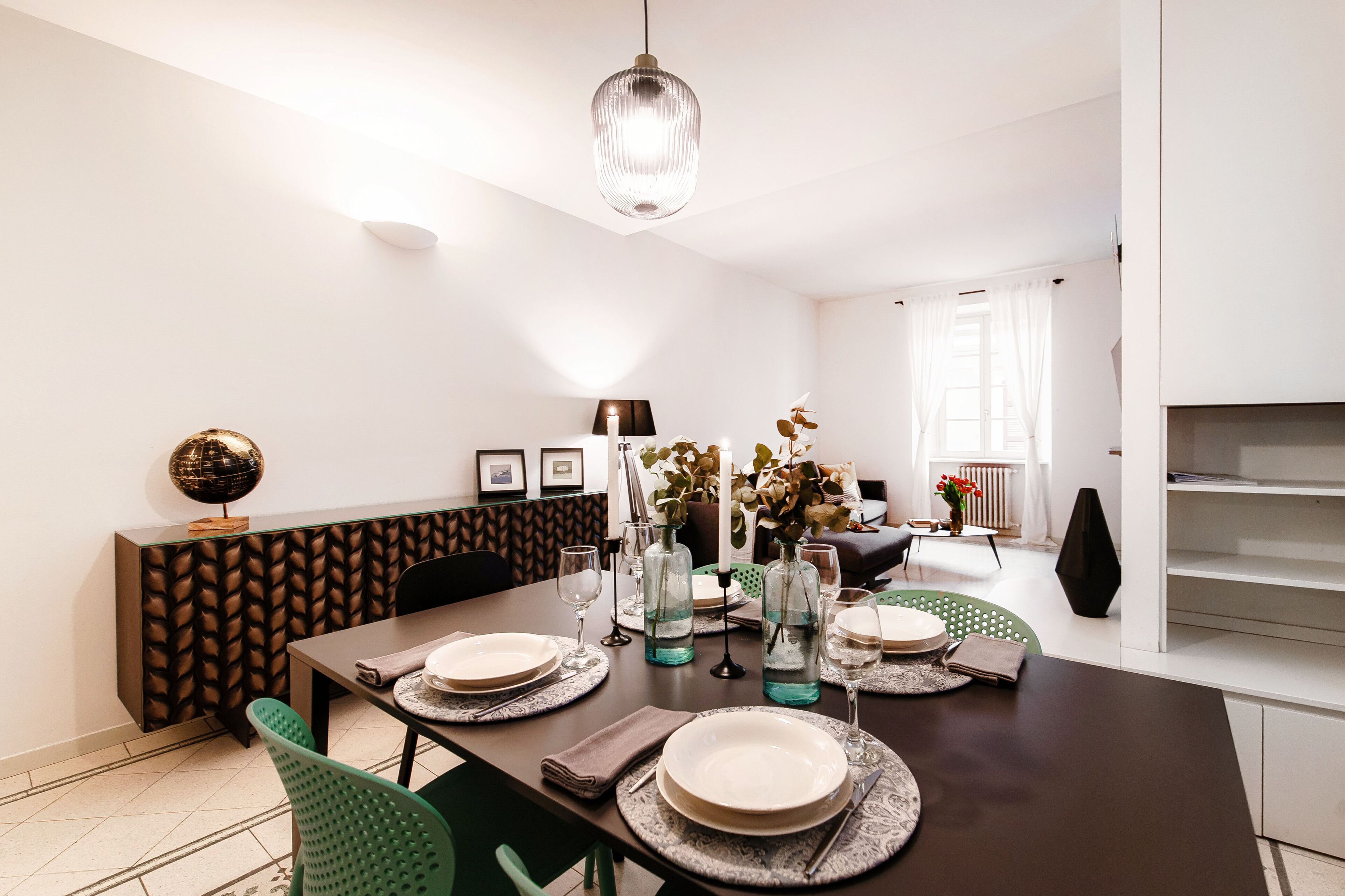 A dining area with a dark wooden table set for four, surrounded by green chairs. A decorative globe is on a cabinet, and a vase with flowers is on the table.